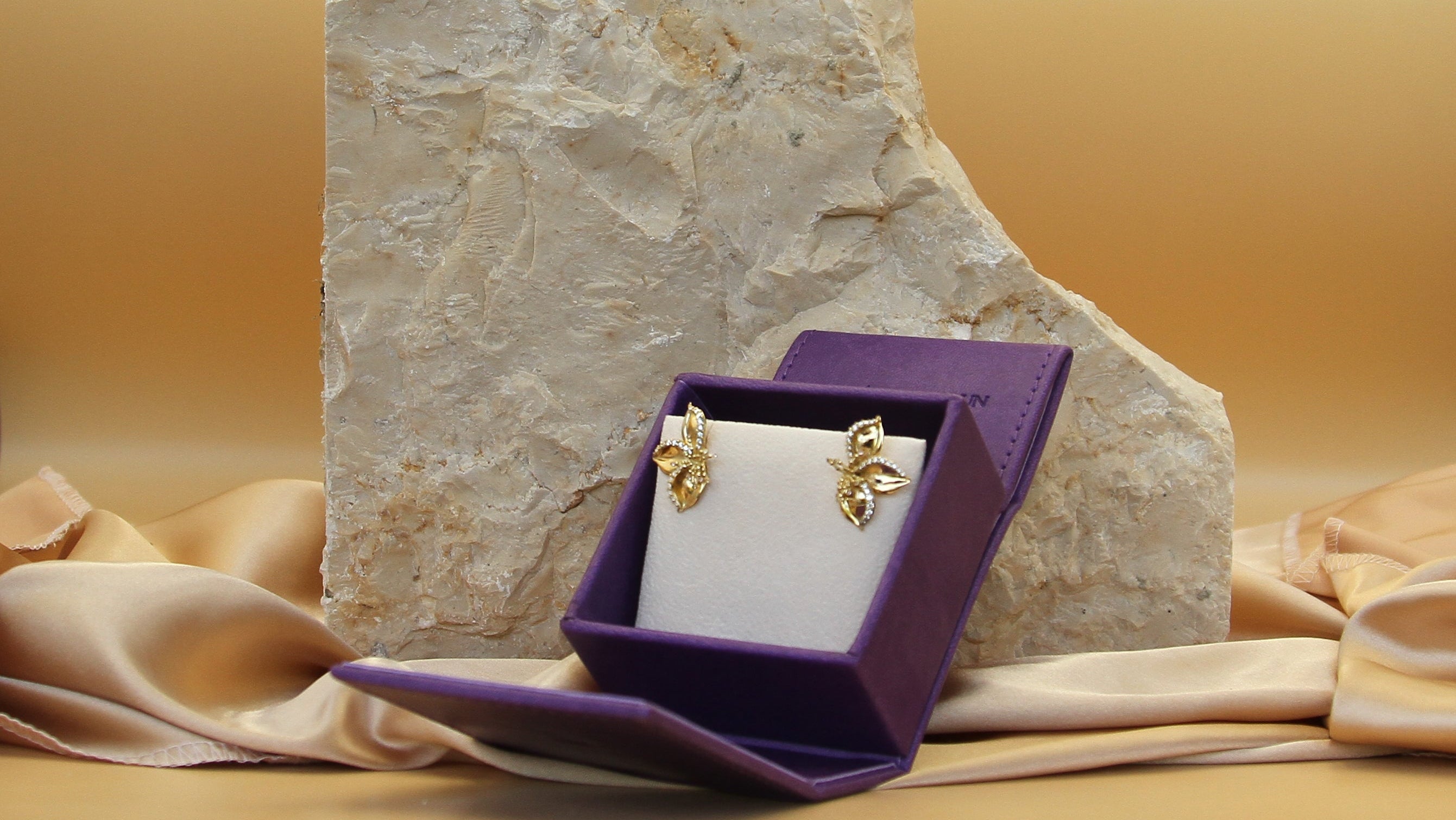 Frangipani earrings designed by Renée Chamoun placed in a purple jewelry box with a stone and fabric background